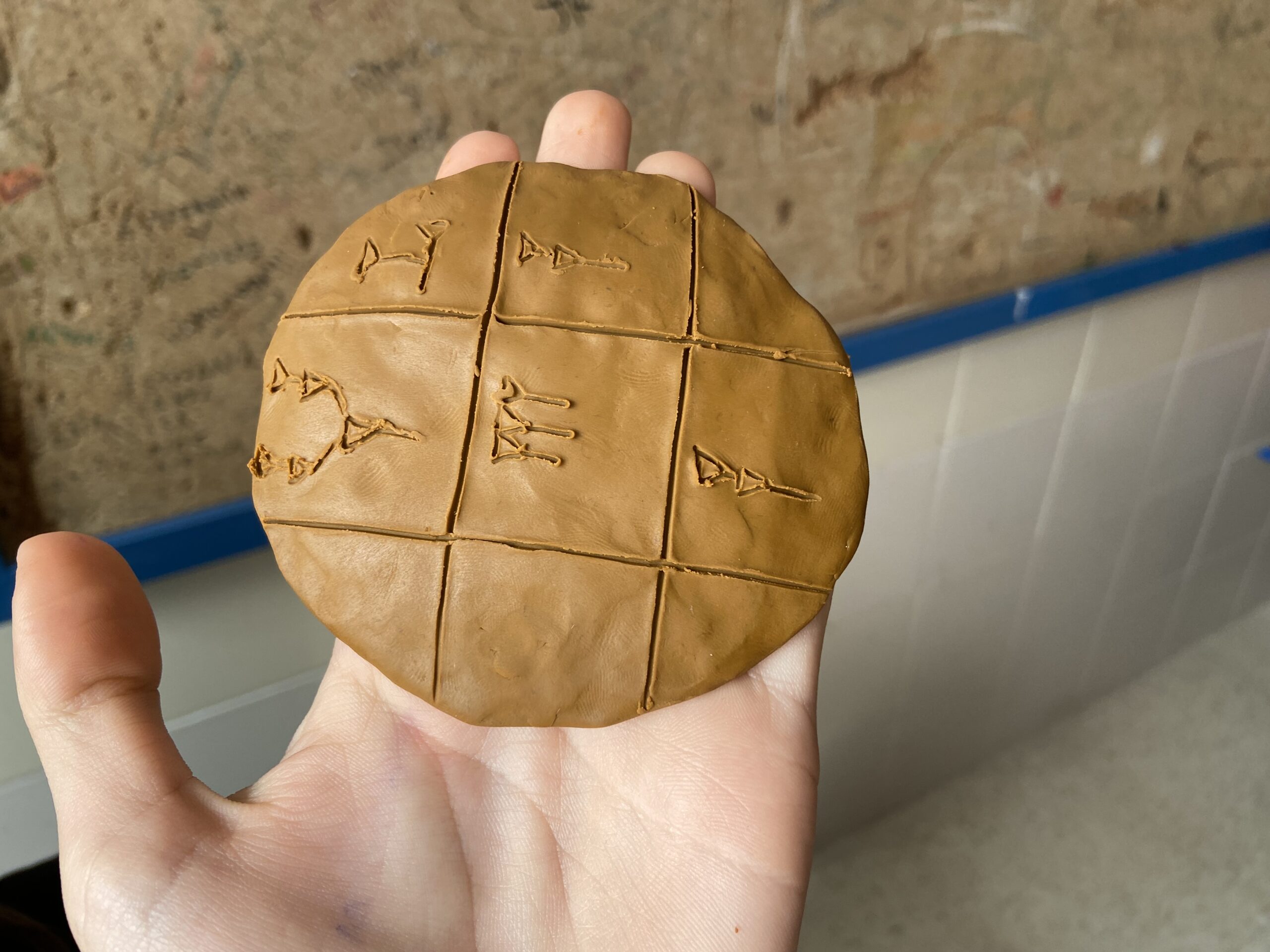 Una mano (la mía) sostiene una tablilla de escritura cuneiforme hecha en plastilina. De fondo, un corcho que ha conocido tiempos mejores.